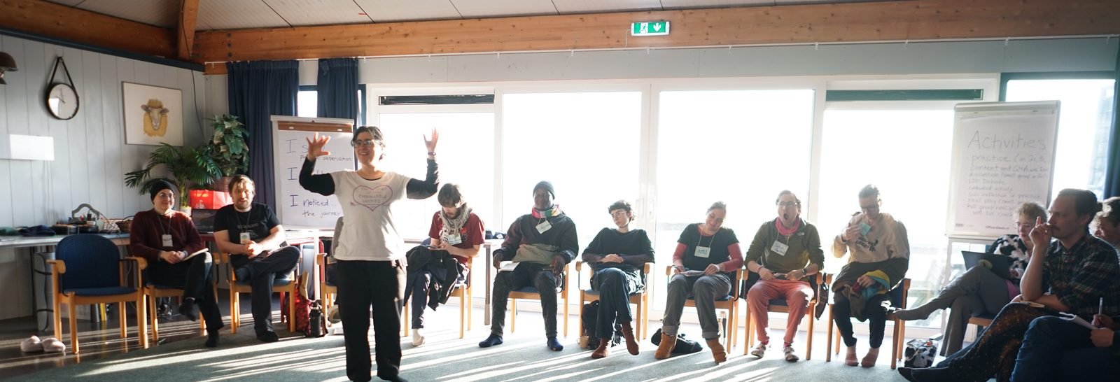 A facilitator stands with open arms in the middle of a circle of seated participants during a sunlit workshop